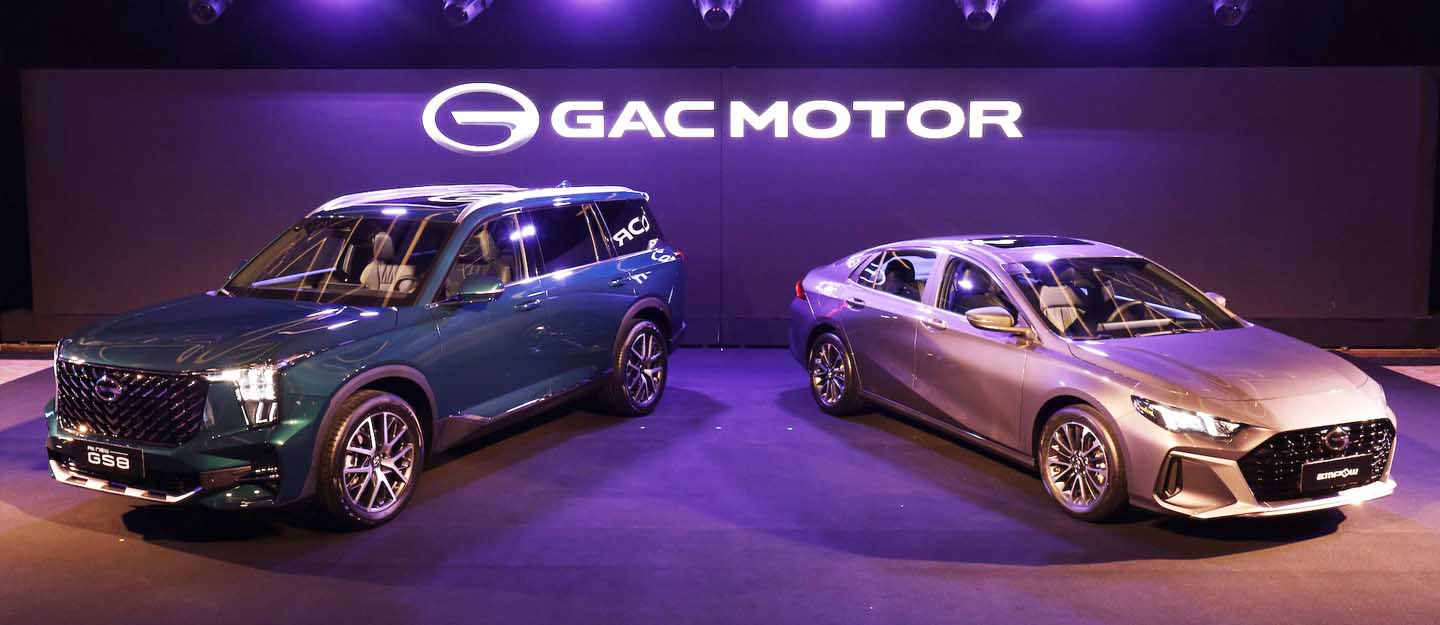 A green GAC GS8 SUV and a silver GAC Empow sedan are displayed on a stage with a "GAC MOTOR" illuminated sign in the background, showcasing GAC car models in the UAE.