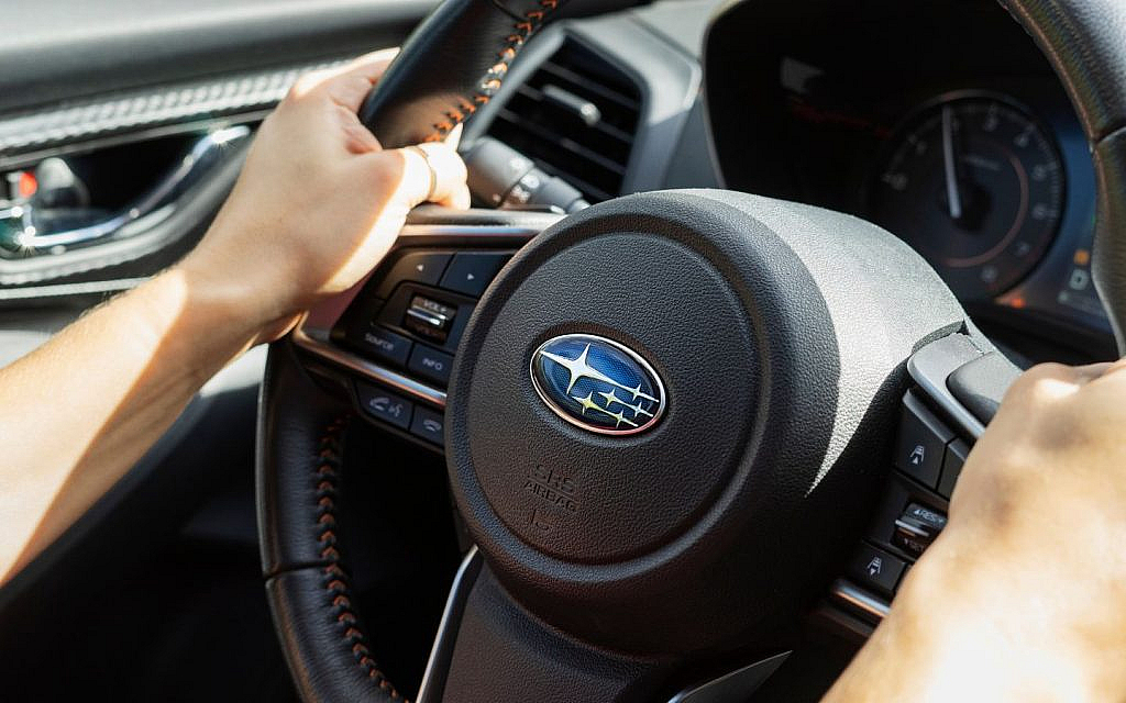 Hands gripping the steering wheel of a Subaru car, prominently displaying the blue and silver six-star emblem in the center.