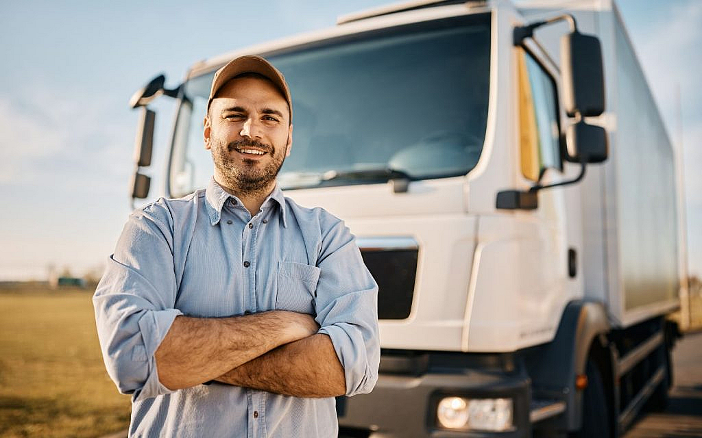 A truck driver with arms folded, standing confidently in front of a heavy vehicle.