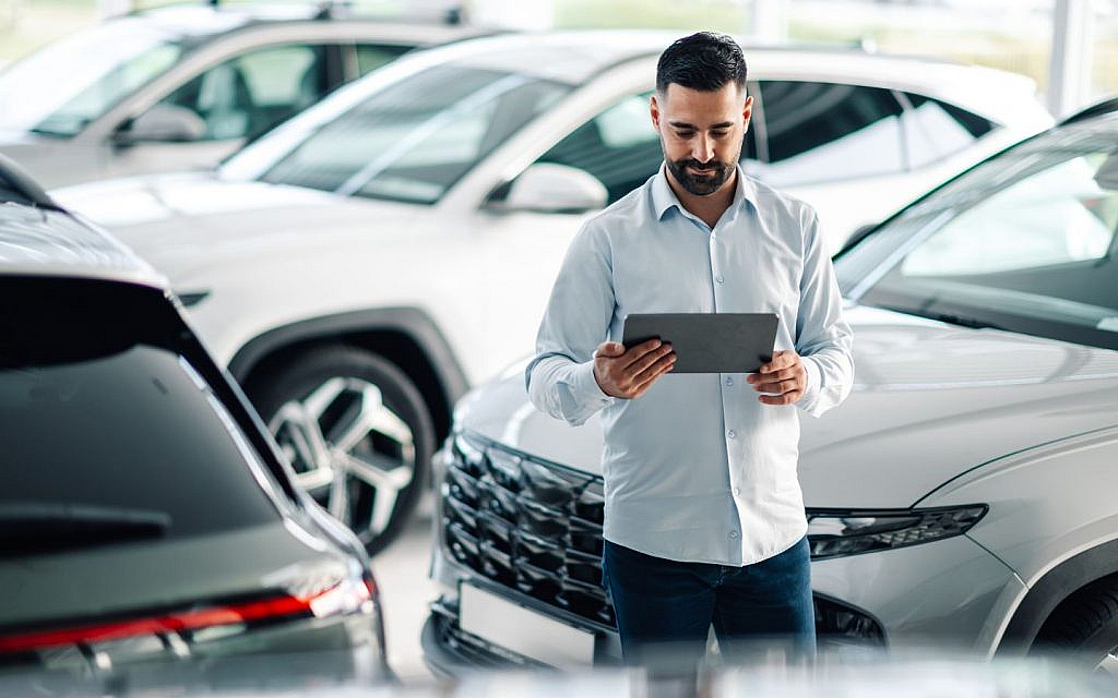 A man in a car dealership is focused on a tablet, with vehicles displayed in the background.