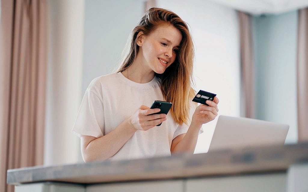 Woman sitting on a chair in front of a small table with an open laptop, holding a phone in one hand and a credit card in the other. 