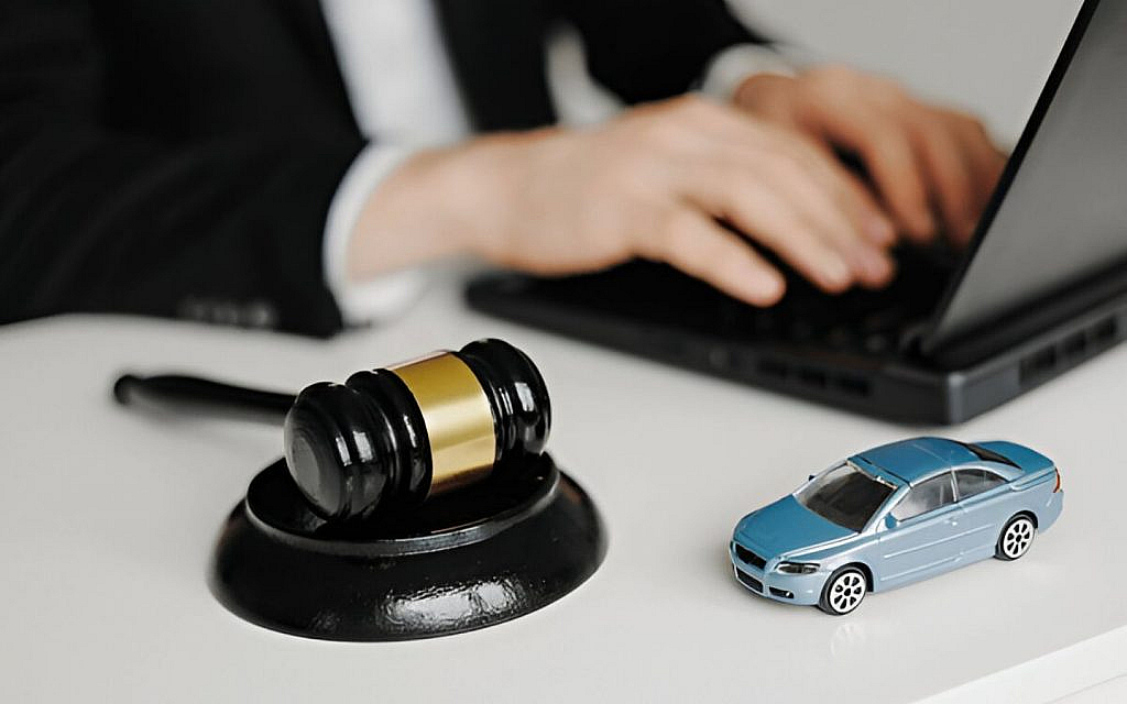 Close-up of a miniature blue car and auction gavel on a white desk, with a person in a suit typing on a laptop in the background