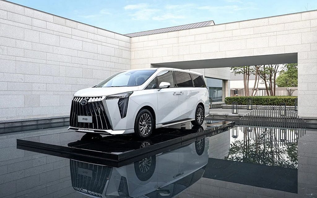 A white GAC M8 minivan with a large, striking grille and chrome accents is parked on a glossy black platform, surrounded by a shallow pool of water that reflects the vehicle. 