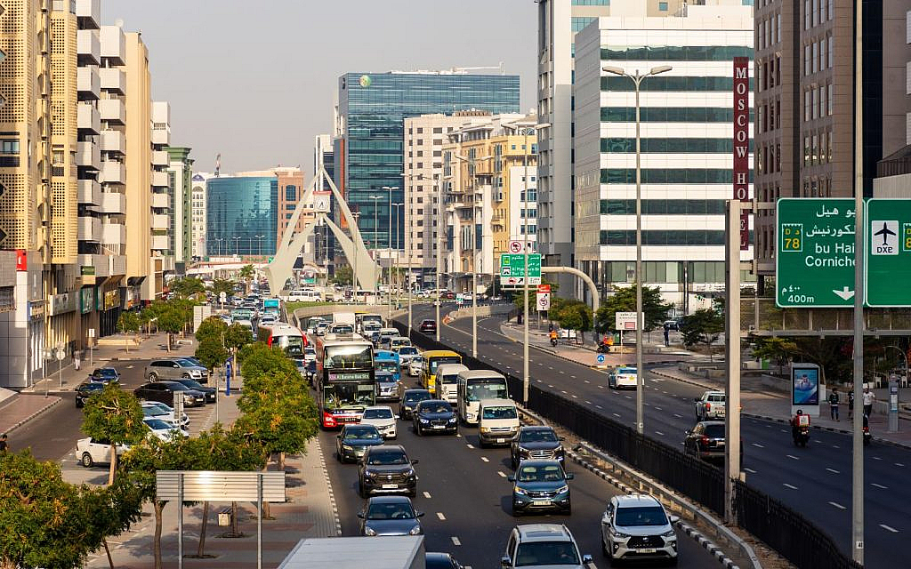 A busy multi-lane city road with cars and buses, lined with modern buildings and street signs.
