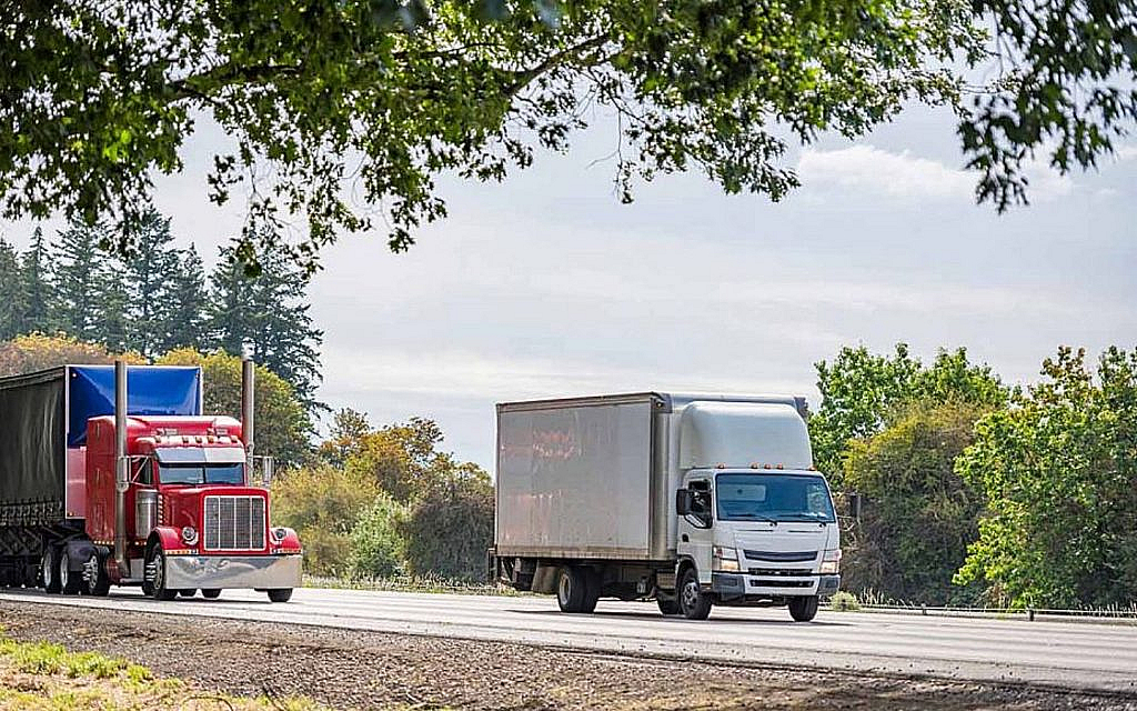 A red semi-truck and a white box truck are driving on a highway with trees alongside the road.
