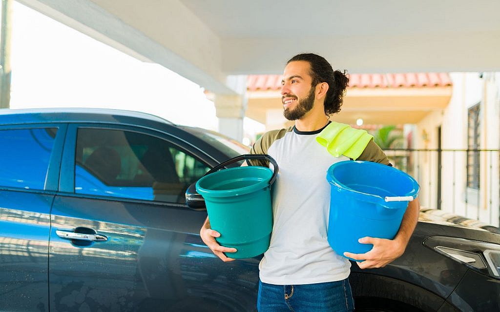 A man with a microfiber cloth on his shoulder, holding two buckets, standing next to a car.