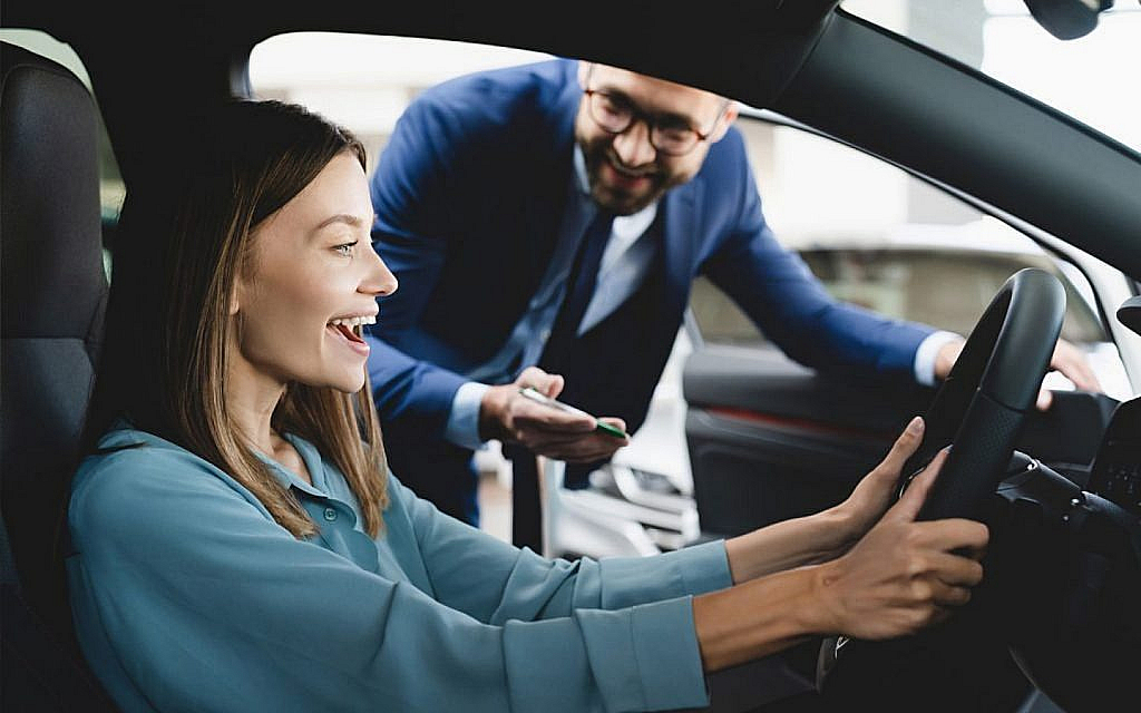 Smiling woman sitting in the driver's seat of a car, with a man in a suit smiling outside the car door.
