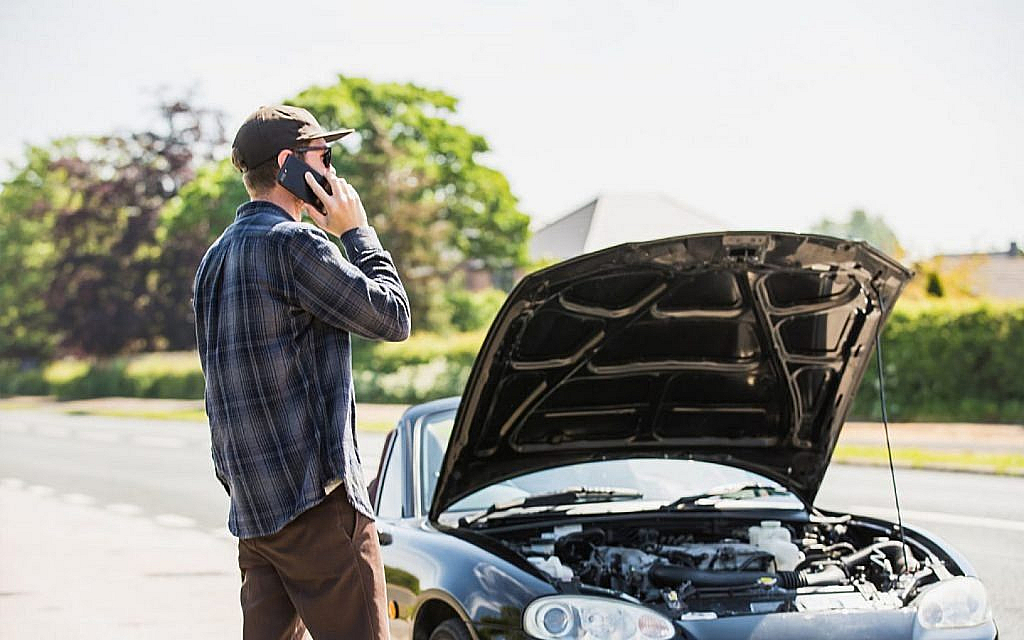 Man in a baseball cap talking on a phone, standing next to a car with its hood open on the side of a road.