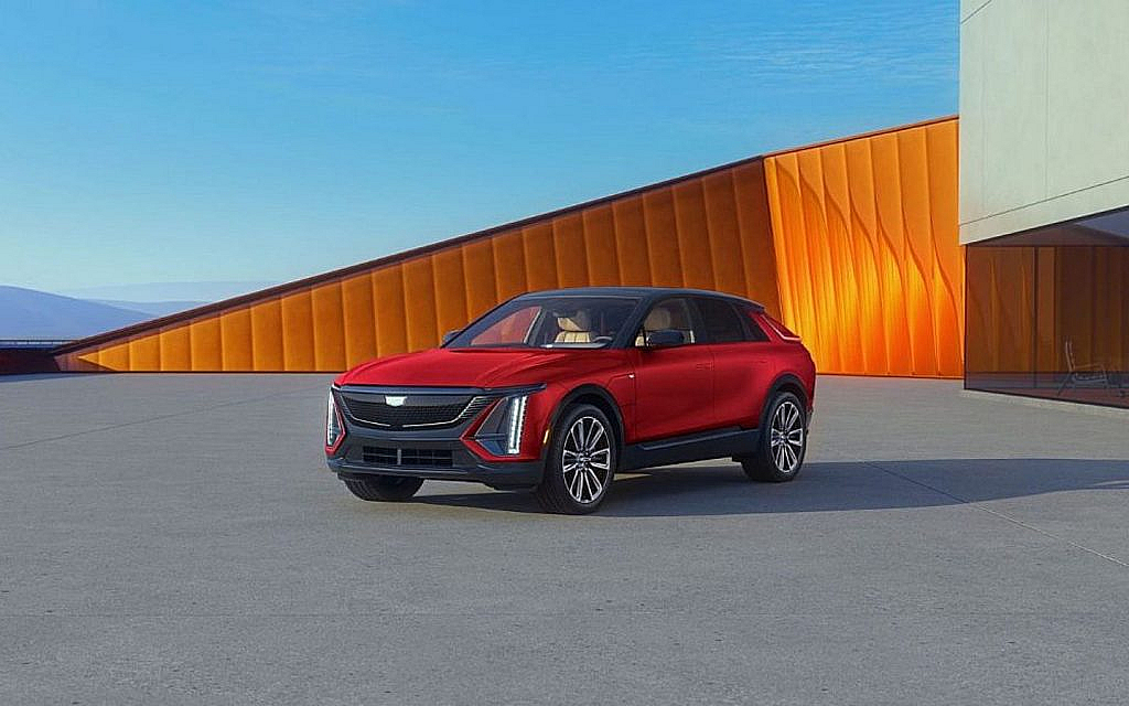A red 2025 Cadillac Lyriq electric SUV is parked on a concrete lot in front of a building with orange and gray walls.