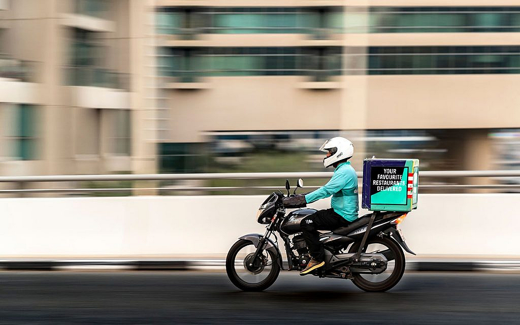 A delivery rider on a motorcycle with a branded box, moving past a building in a city.