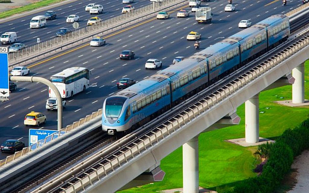A modern public transit train on an elevated track running parallel to a multi-lane highway filled with traffic.