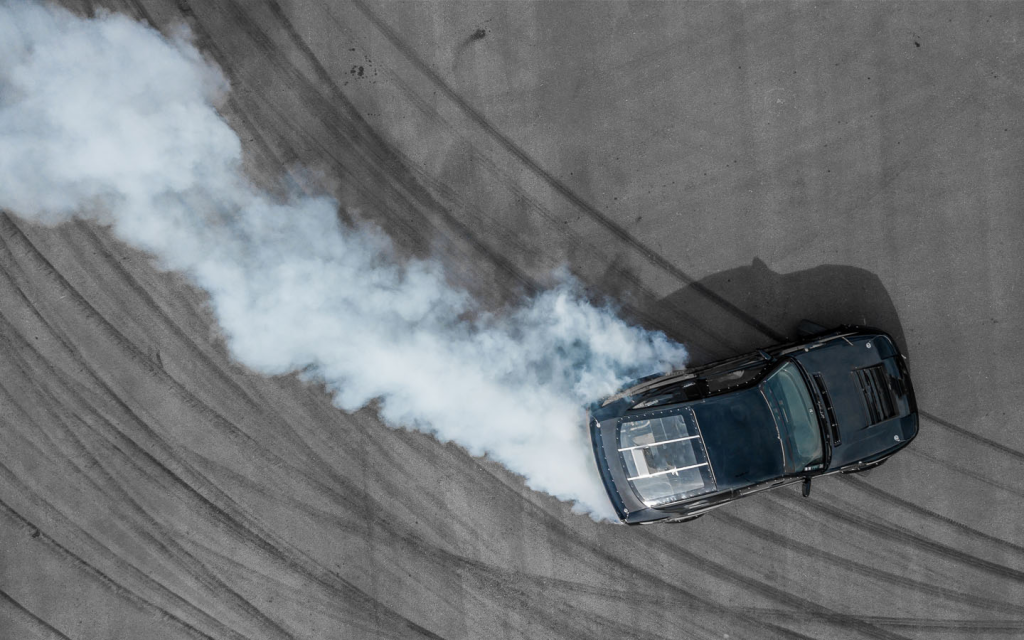An aerial high-angle shot captures a dark sports car drifting. 