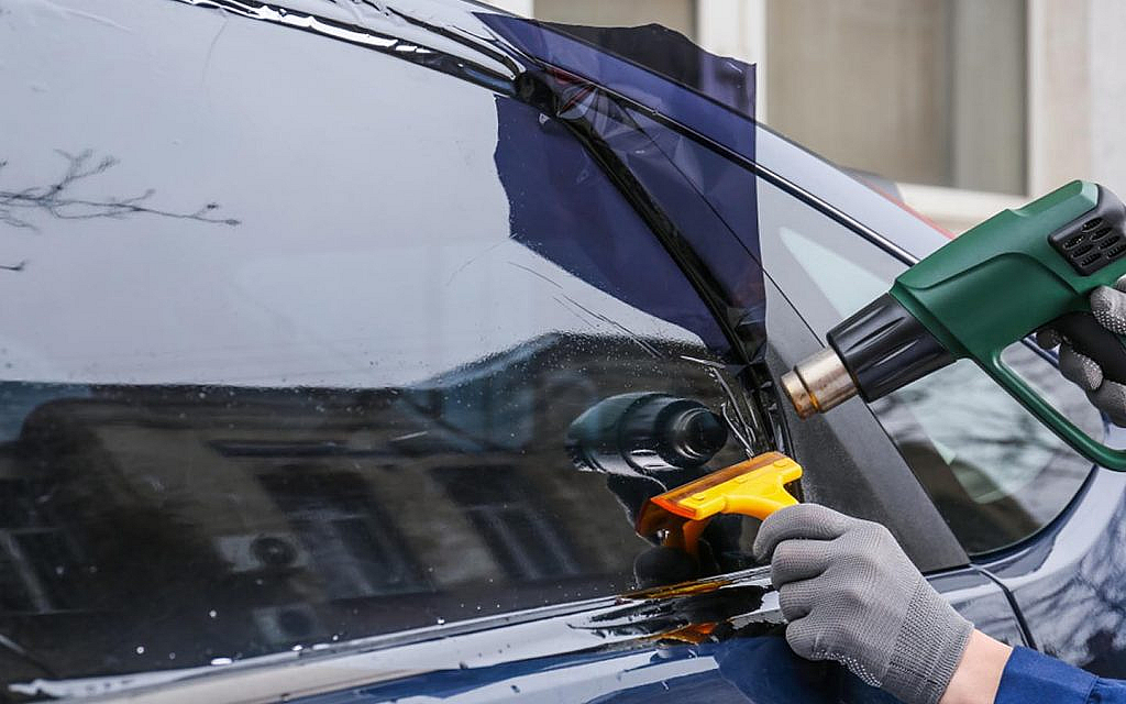 A professional uses a heat gun and a yellow squeegee to shrink and smooth a dark tint film. 