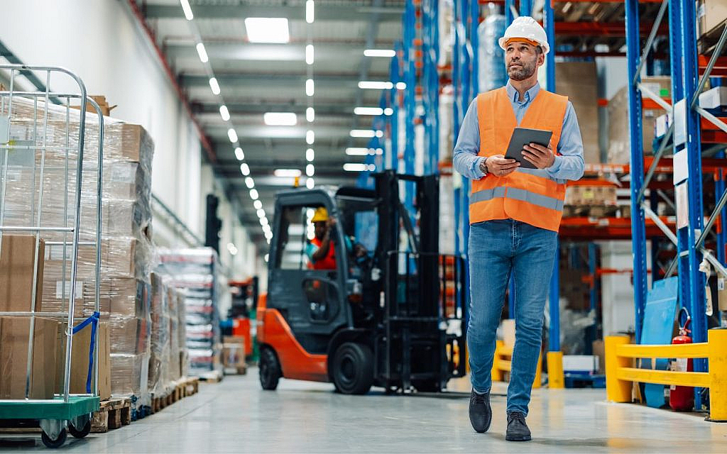 A male supervisor in a safety vest and hard hat holds a tablet while walking through a busy warehouse.